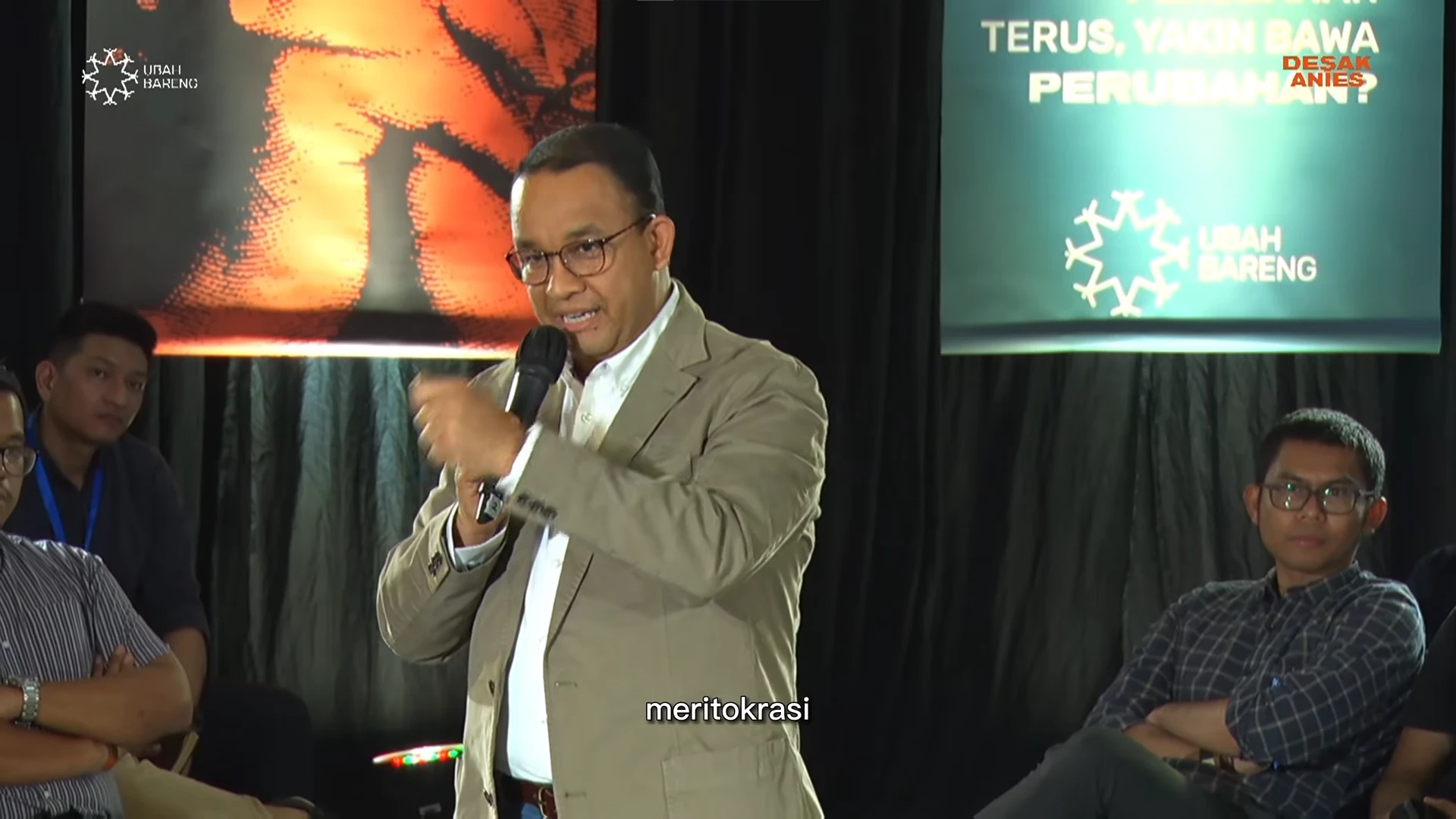 Anies stands with a microphone answering a question among seated audience members, with Desak Anies and Ubah Bareng banners behind him
