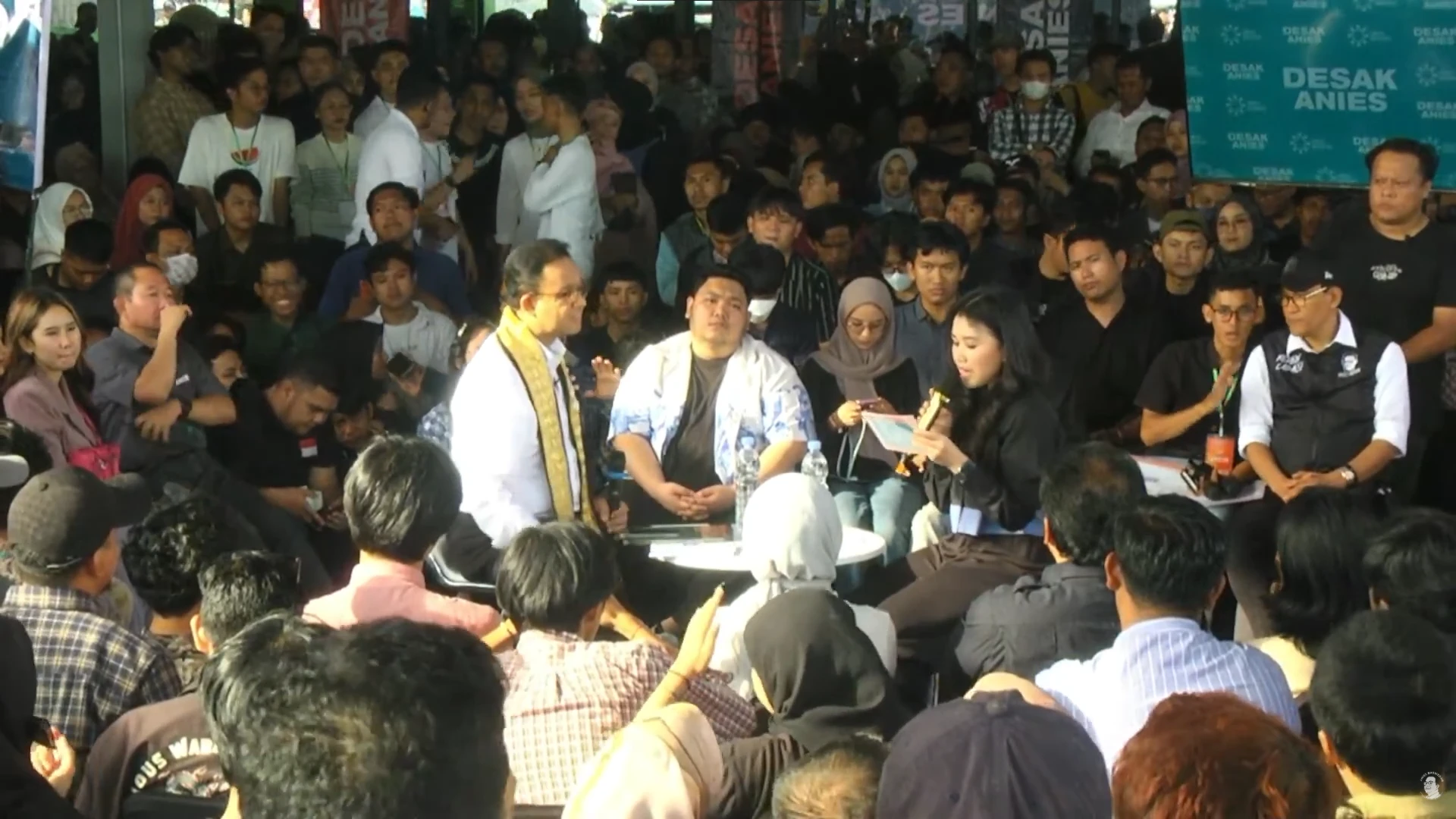 An open-air session: Anies sits at a small table in the middle of a dense crowd of young people while the moderator reads out a question, in front of a Desak Anies backdrop