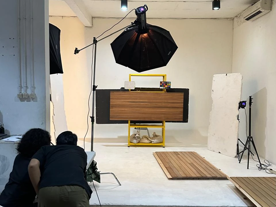 A photo studio with a white cove: a wooden deck panel mounted upright on a yellow stand under a large softbox, with a colour checker and a label card above it, more boards laid on the floor, and two people bent over a laptop in the foreground